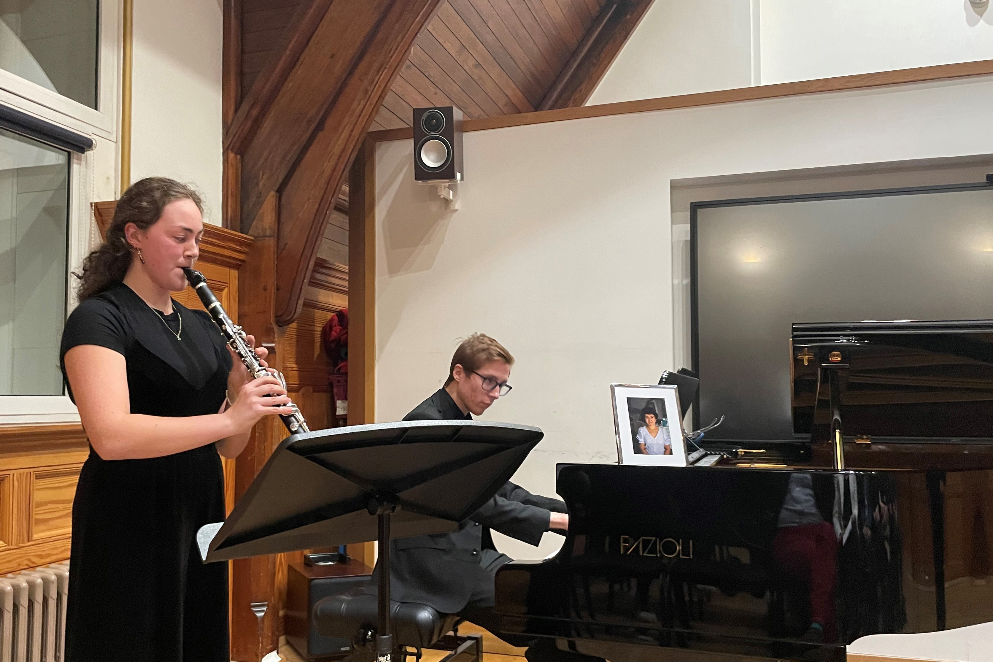 A woman wearing a dark black dress, playing the clarinet, with a man, wearing smart black attire, playing the piano in a wood panelled room.